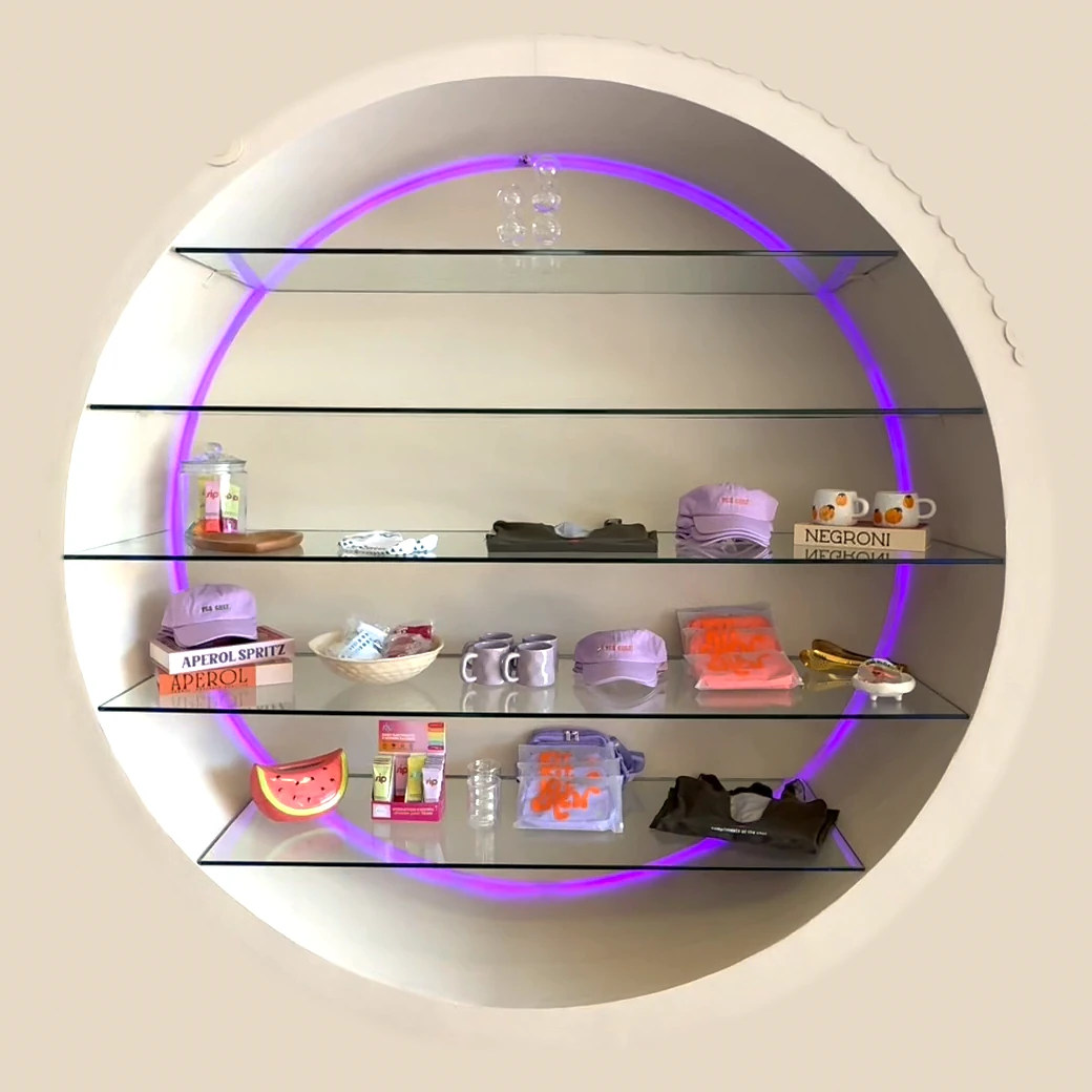 The merch circle at The Kitchen: a round wall niche with four glass shelves of caps, mugs, grip socks, tees and bags under a lilac neon ring