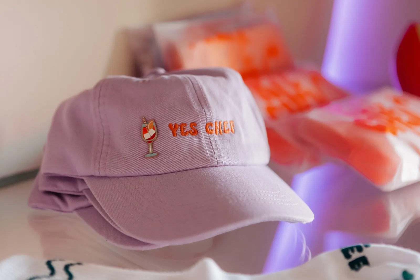 The Kitchen’s lilac “yes chef” cap, with its spritz pin, on the glass shelf of the studio’s merch circle